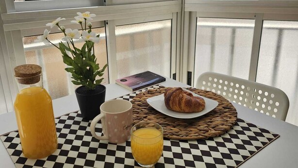 Et morgenbord dækket med et ternet borddække, appelsinjuice, en kop, en tallerken med croissanter, en bog og en potteplante.