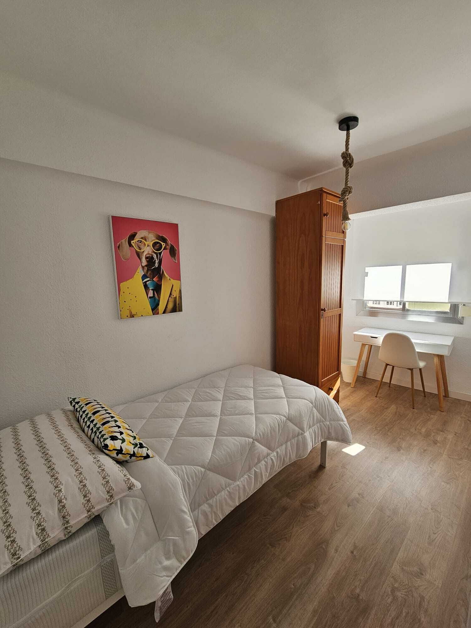 A bedroom with a bed, a desk, and a chair. There is a painting on the wall and a wooden wardrobe.