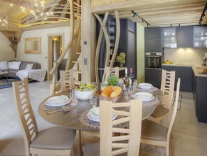 Uma sala de jantar com uma mesa redonda posta para quatro pessoas, rodeada por cadeiras de madeira. Uma mesa de buffet com salada e pão está no centro. O espaço tem um design moderno com uma escada que leva a um andar superior.
