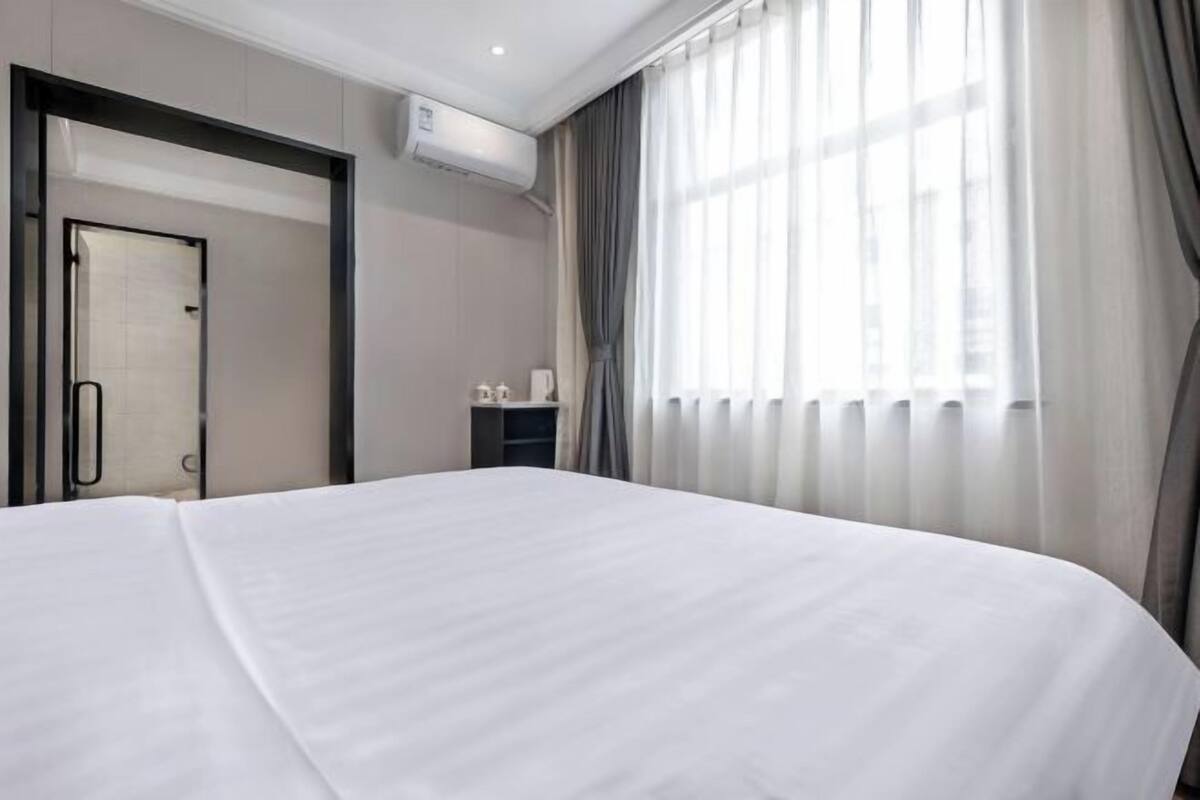 Ein Hotelzimmer mit einem großen Bett, einer Tür, einem kleinen Tisch mit einer Tasse und einem Fenster mit Vorhängen.