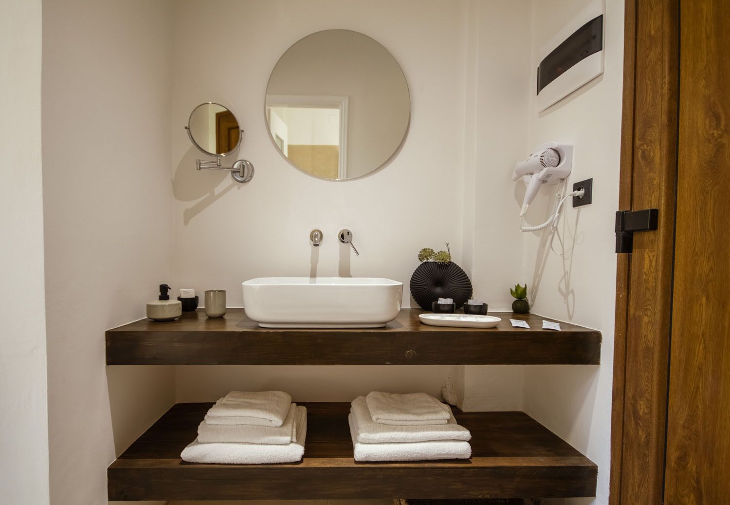 A modern bathroom with a white sink, two faucets, a round mirror, a hairdryer, and neatly folded towels on a wooden shelf.