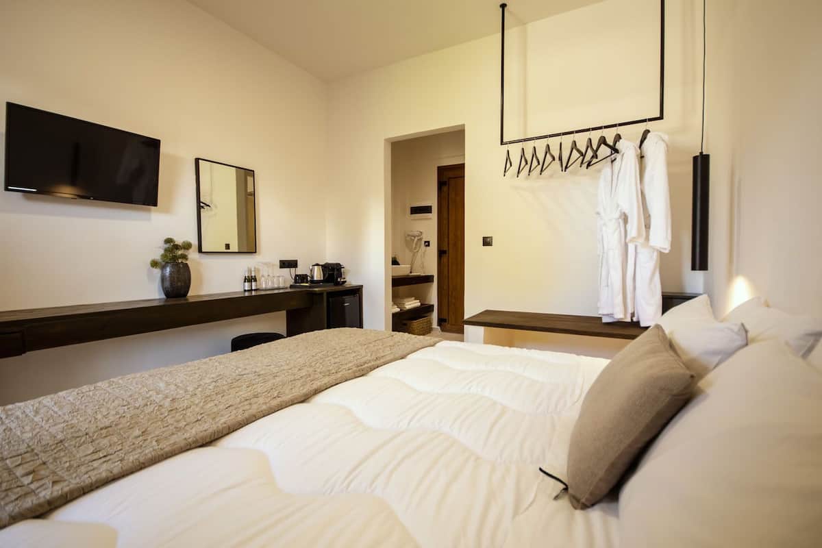 A hotel room with a large bed, a desk with a coffee machine, a TV, and a mirror. There is a robe rack with towels and a door leading to a bathroom.