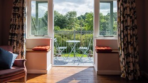 Ein Zimmer mit Holzboden, einem rosa Sofa, einem kleinen Tisch mit Stühlen und einem Balkon mit Blick auf Grünflächen.