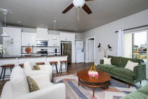 Una cocina moderna con armarios blancos, una isla central y taburetes de bar. En la zona de estar hay un sofá verde y un sillón blanco, con una mesa de centro en medio.