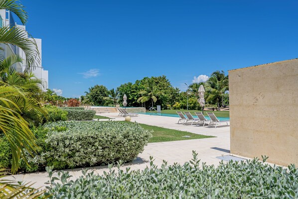 A pool area with lounge chairs, a palm tree, and a building in the background. - Cozy 1-bedroom condo with direct pool access, AC & Wi-Fi in Pleasant Punta Cana (Punta Cana)