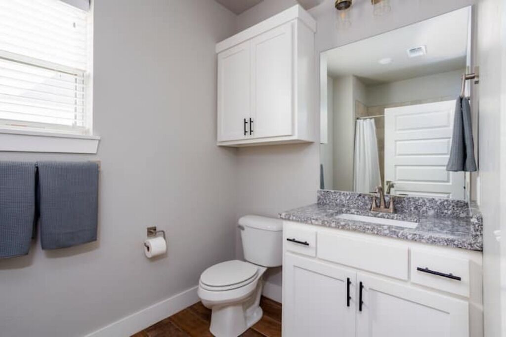 Une salle de bain avec des meubles blancs, des toilettes, un lavabo doté d’un plan de travail en marbre et un miroir au-dessus du lavabo.