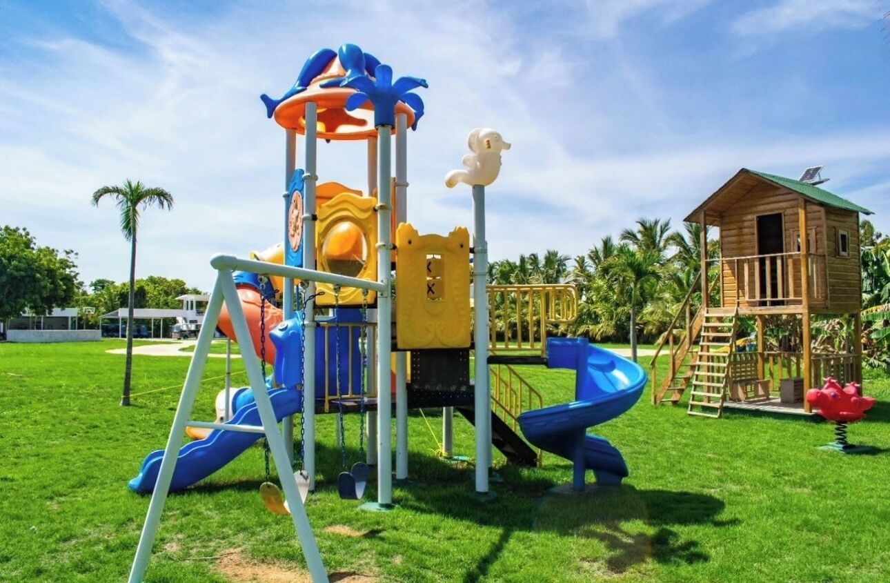 Un parque infantil con toboganes y estructuras para escalar, rodeado de césped y un edificio de madera.