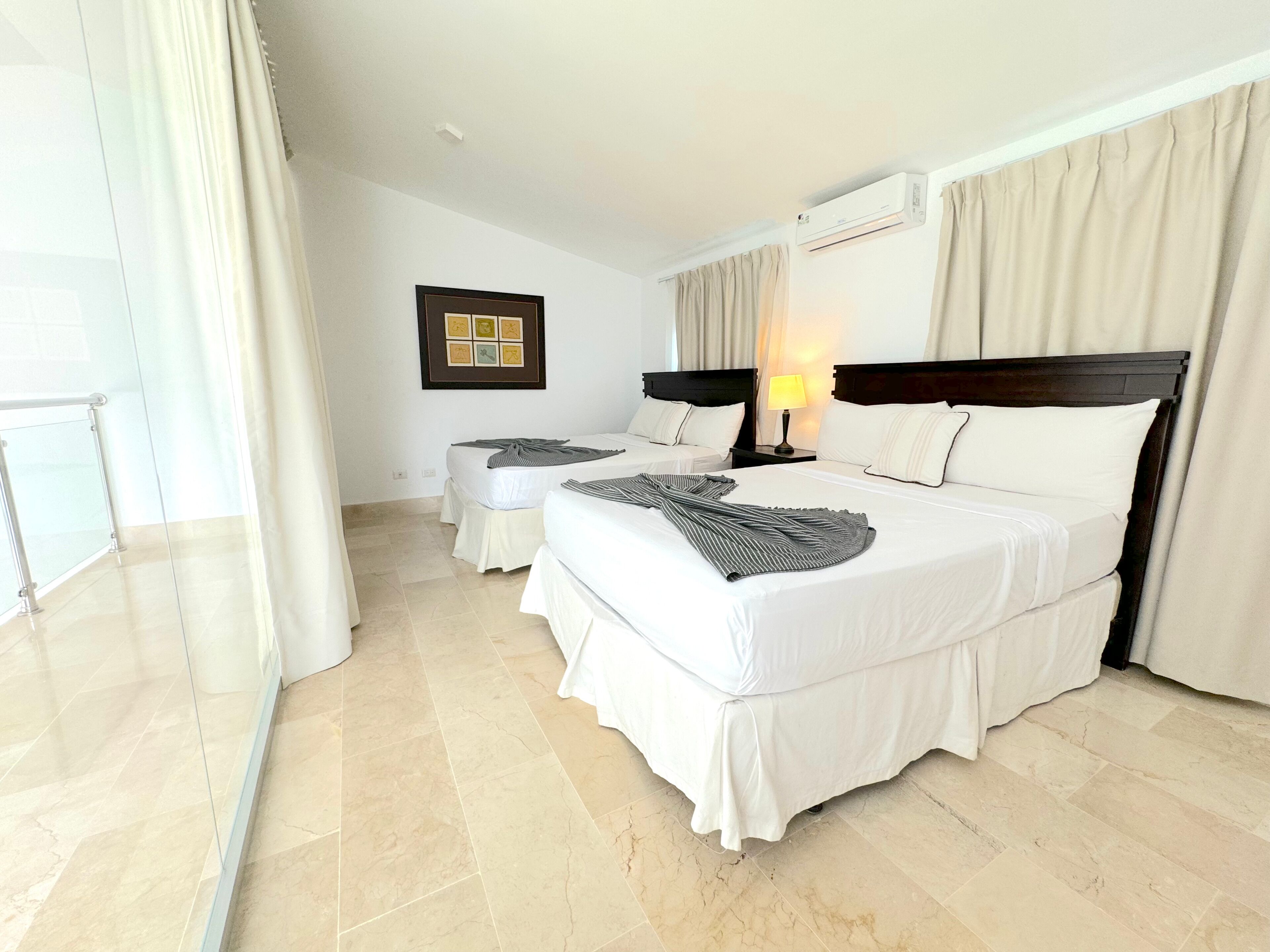 A hotel room with two beds, a large window with curtains, an air conditioning unit, and a framed picture on the wall.
