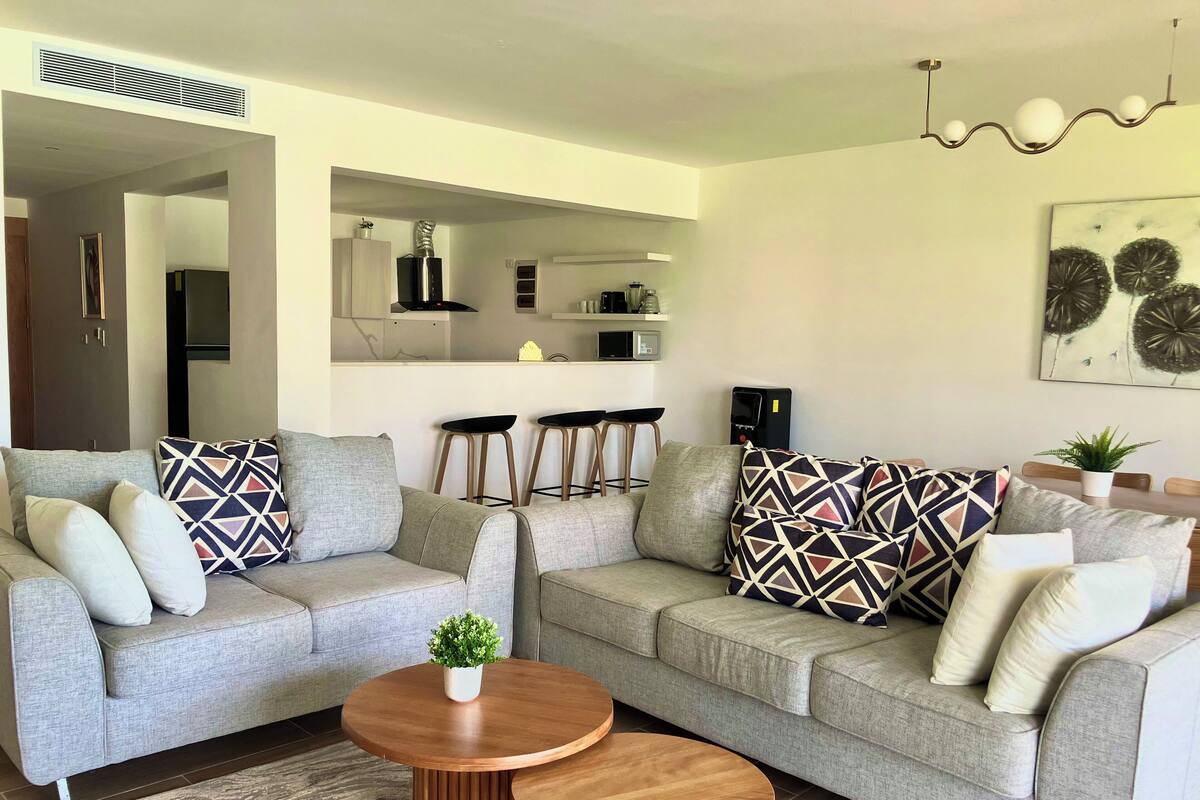 Ein modernes Wohnzimmer mit einem grauen Ecksofa, zwei Holztischen und einer kleinen Topfpflanze.