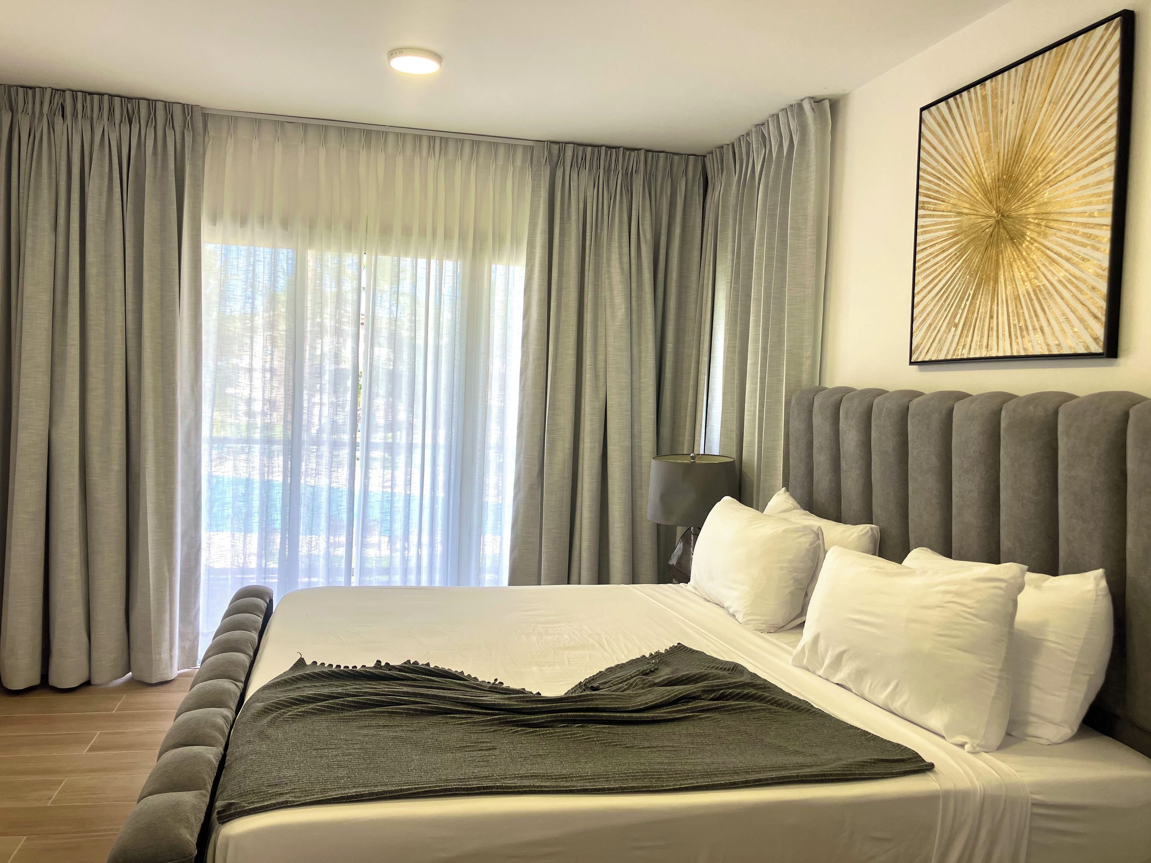 A modern bedroom with a large bed, grey curtains, a wall-mounted lamp, and a decorative piece above the bed.