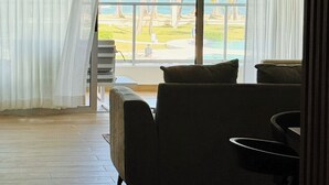 A room with a large window overlooking a beach and palm trees. There is a sofa and a small table in the room. - Playa Palmera Beach Front Apartments (Punta Cana)