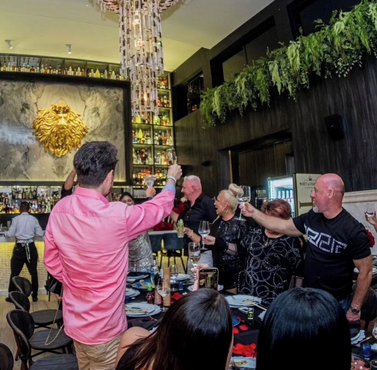 A bar area with a high ceiling, a decorative chandelier, and a wall of shelves with bottles.