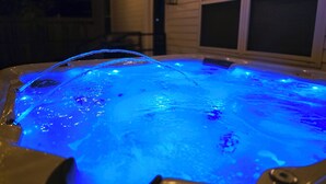 A hot tub with blue lighting, surrounded by a house with a porch and string lights.