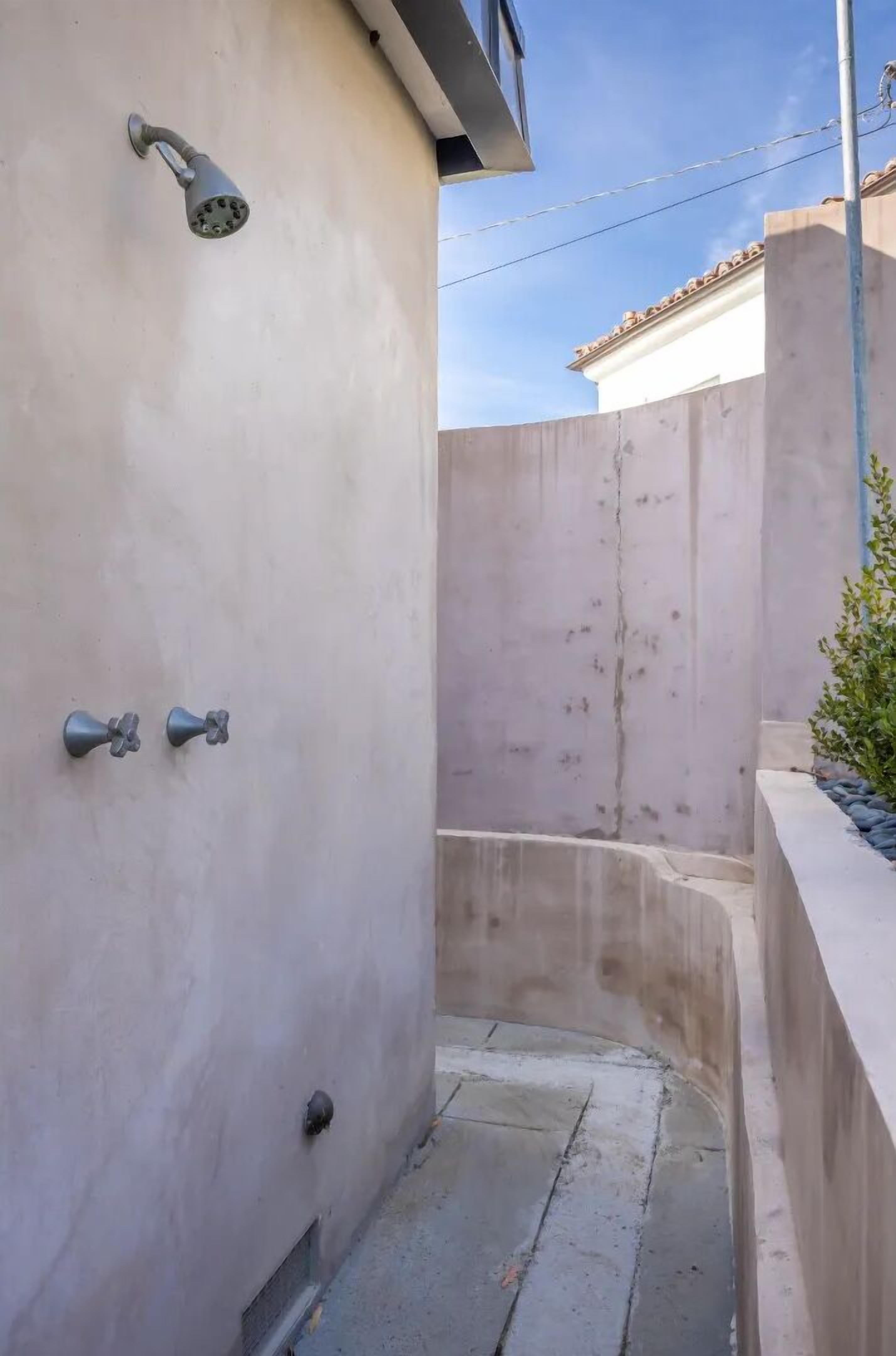 Un étroit chemin de béton mène à un espace douche équipé d’une pomme de douche fixée au mur.