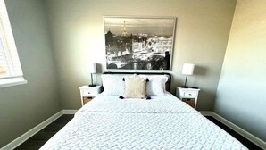 Un lit bien fait, recouvert d’une courtepointe matelassée, deux tables de chevet avec des lampes, et un grand tableau représentant un paysage urbain accroché au mur.