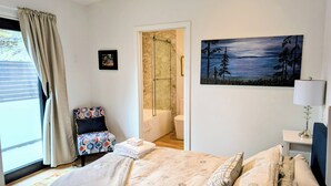 A bedroom with a bed, a chair, a painting, a bathroom with a shower, and a window with curtains. - New Oceanfront Luxury Suite with a breathtaking  view of downtown Vancouver, (Bowen Island)