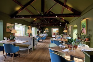 Un restaurante con vigas de madera, paredes verdes y sillas azules. Las mesas están preparadas con platos, vasos y cubiertos.