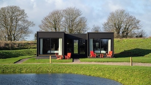 Una casa moderna con amplios ventanales, un pequeño espacio al aire libre con sillas y un estanque en primer plano.