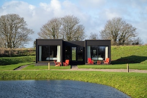 Una casa moderna con amplios ventanales, un pequeño espacio al aire libre con sillas y un estanque en primer plano.