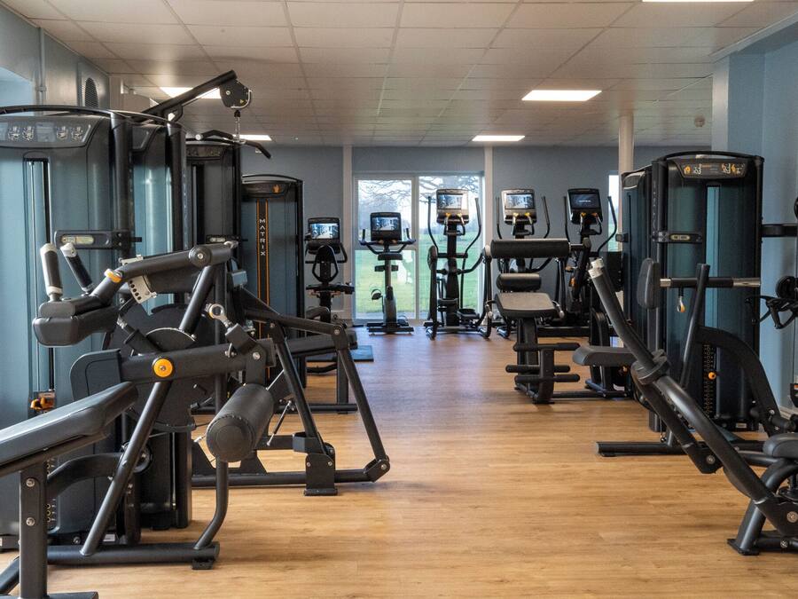 Un gimnasio con diversas máquinas de ejercicio, suelo de madera y grandes ventanales que permiten la entrada de luz natural.