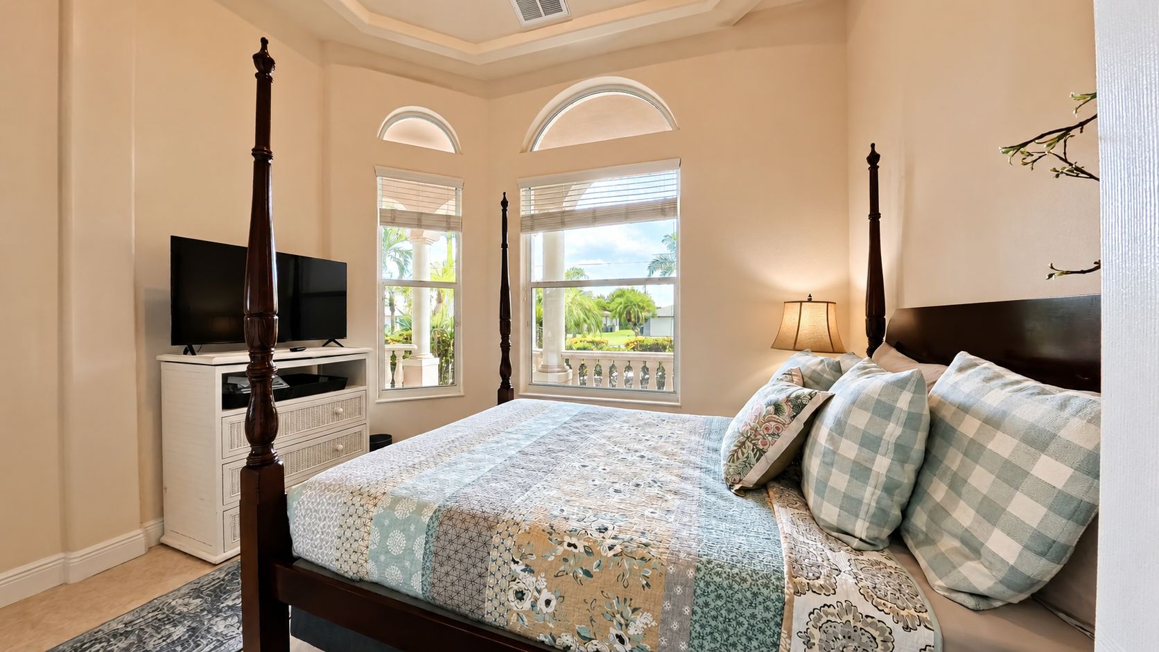 A bedroom with a four-poster bed, a television on a dresser, and a balcony with a view of greenery.