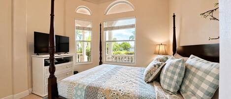 A bedroom with a four-poster bed, a television on a dresser, and a balcony with a view of greenery.