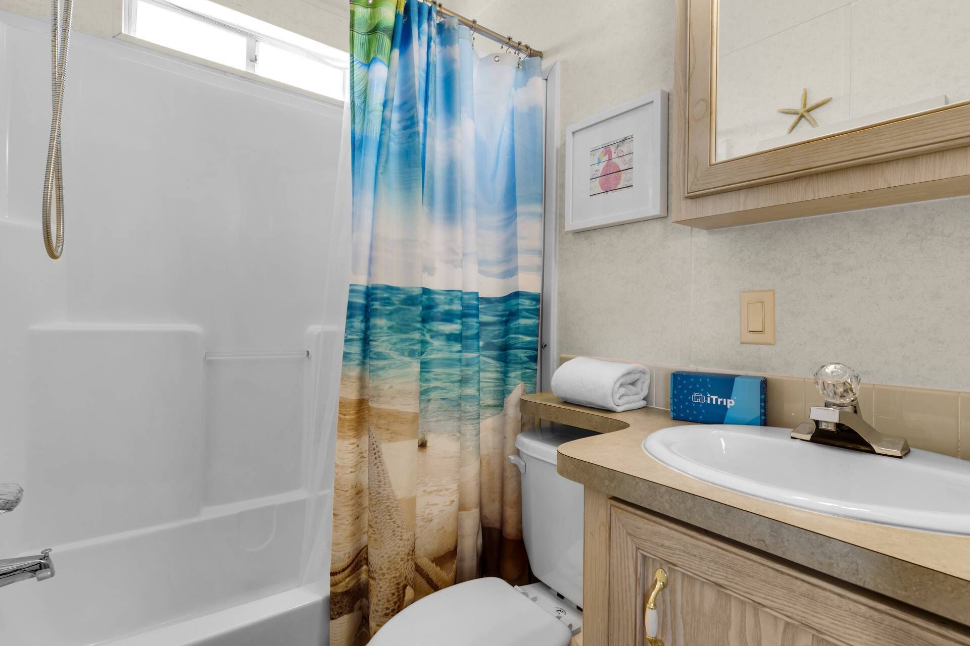 A bathroom with a sea-themed shower curtain, a white toilet, a sink with a blue towel, and a framed picture of a flamingo.