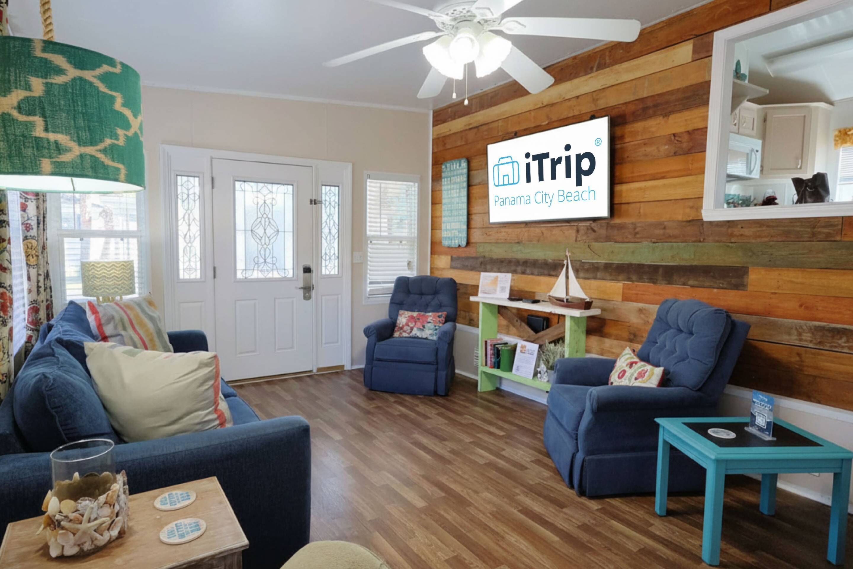 A living room with a wooden wall, a ceiling fan, a sofa, two armchairs, a small table, and a sign that reads 'iTrip Panama City Beach.'