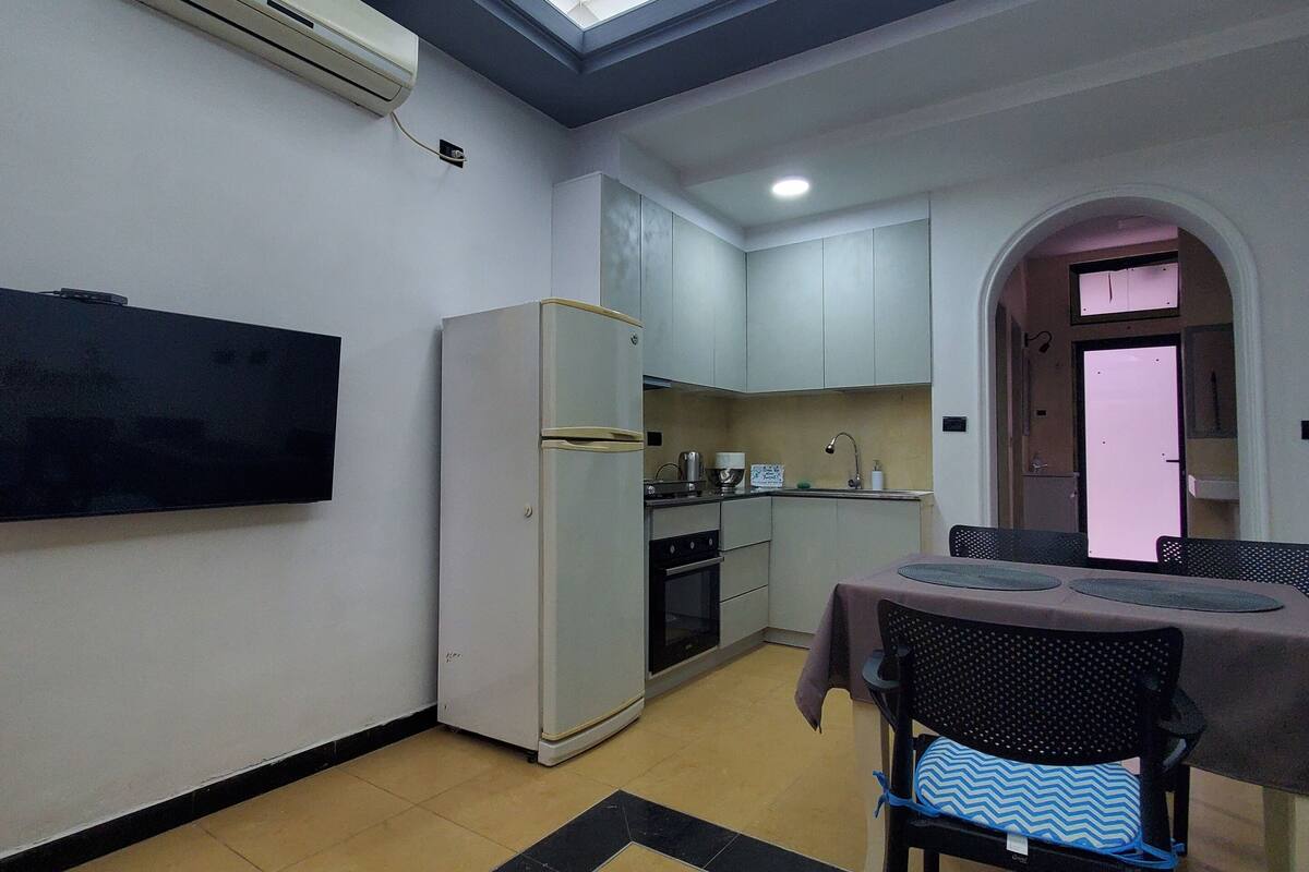 A compact kitchen with a refrigerator, stove, and microwave. There's a dining table with chairs and a ceiling with a unique design.