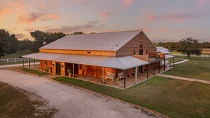 A large wooden barn with a covered porch and a second-story balcony, surrounded by a fenced property.