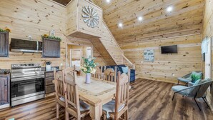 A wooden interior with a dining table, chairs, a kitchen with stainless steel appliances, a clock, and a staircase.