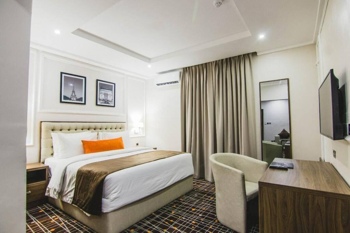 A hotel room with a bed, a desk, a chair, a TV, and two framed pictures on the wall.