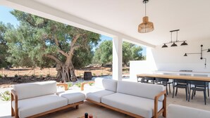 A modern outdoor living area with a white sofa, wooden table, and chairs, overlooking a natural landscape with trees and a clear sky.