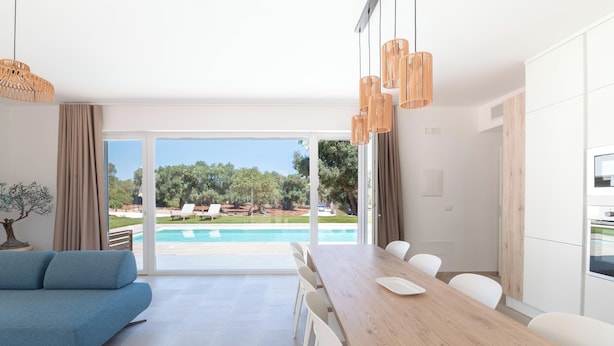 A modern living room with a blue sofa, a wooden dining table, and white chairs. There is a large window with a view of a swimming pool and greenery outside.