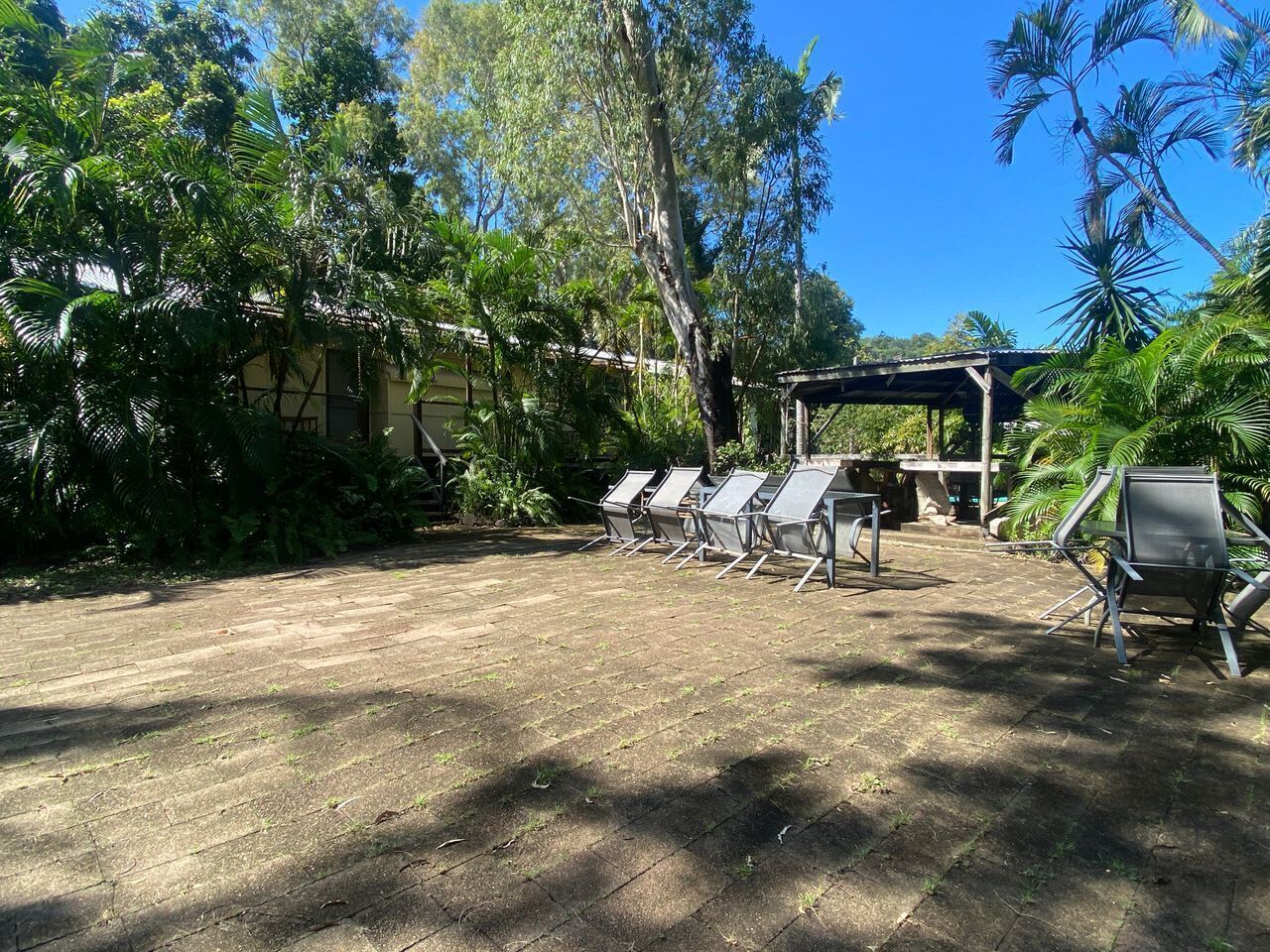 An outdoor seating area with metal chairs and a covered structure, surrounded by lush greenery and trees.