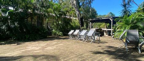 An outdoor seating area with metal chairs and a covered structure, surrounded by lush greenery and trees.