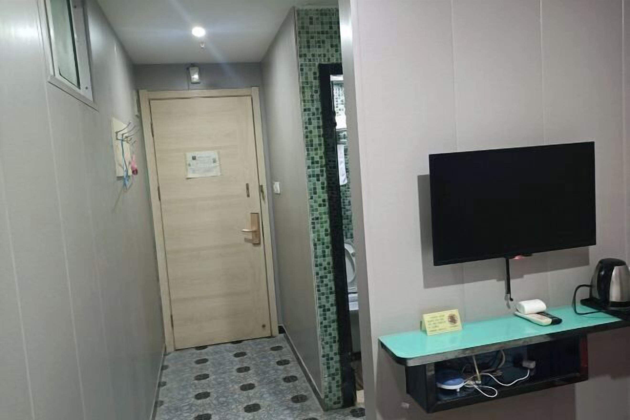 Un corridoio con una porta in legno, una parete piastrellata, un televisore a parete e un piccolo ripiano con un telefono e un bollitore.