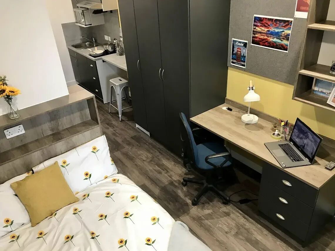 A compact bedroom with a bed, a desk with a laptop, a chair, and a small kitchenette.
