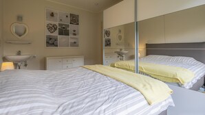 A bedroom with two beds, a large mirror, a bedside table, a dresser, and a wall-mounted shelf with decorative items.
