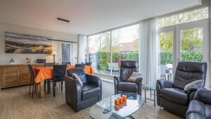 A modern living room with leather sofas, a glass coffee table, and a dining area with orange tablecloths.