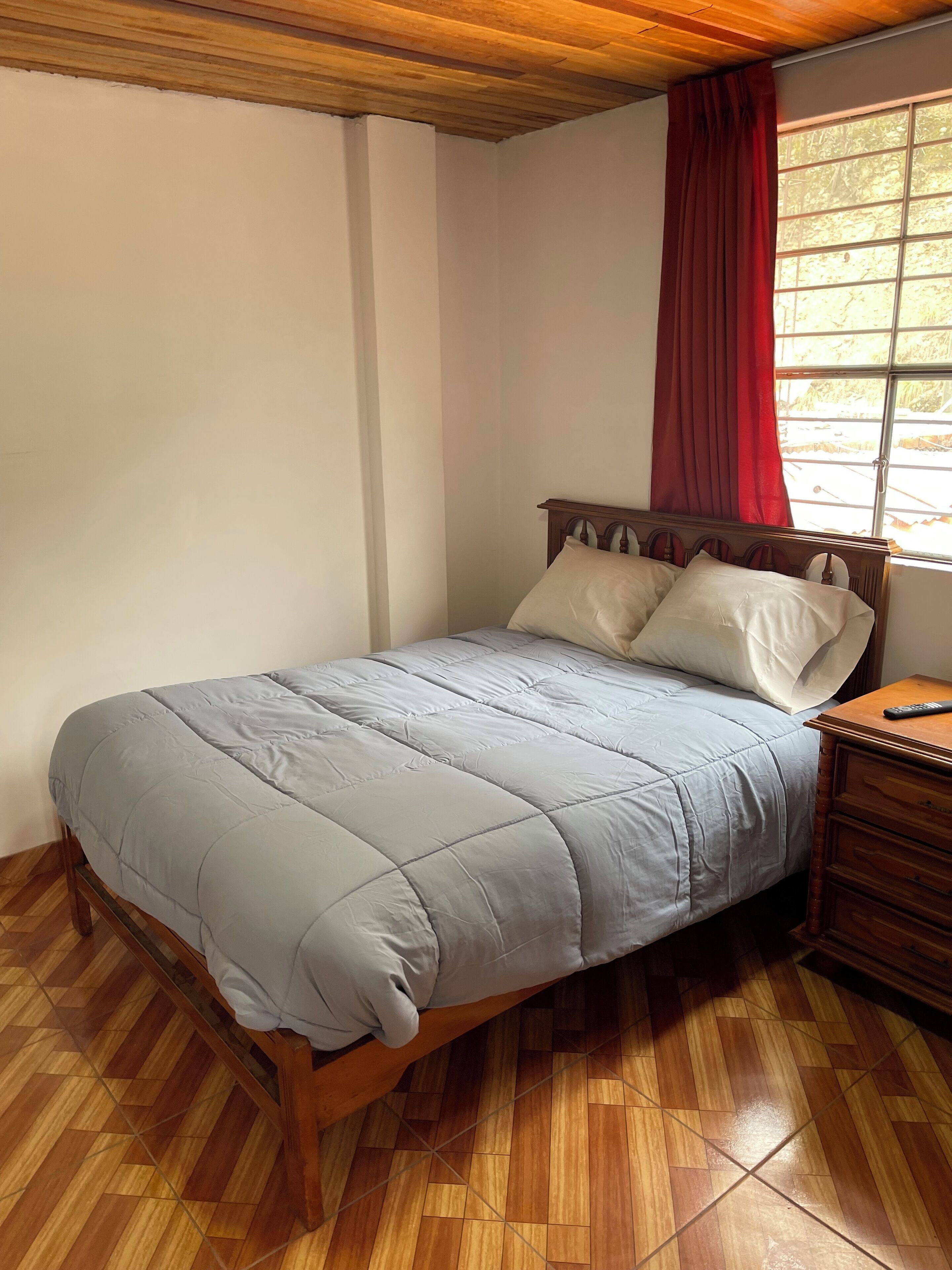 Un lit simple avec une tête de lit en bois, un rideau rouge et une table de chevet en bois avec une télécommande.