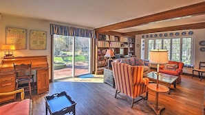 Ein geräumiges Wohnzimmer mit Holzboden, Kamin, Bücherregal und einem großen Fenster mit Blick ins Freie.
