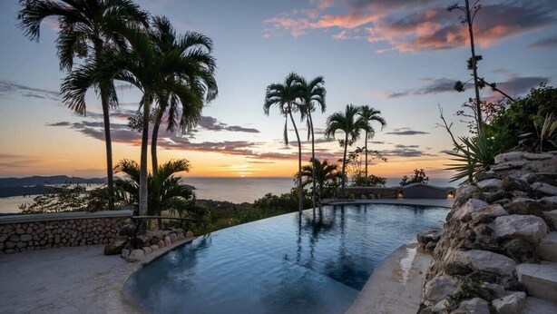 A curved infinity pool with palm trees and a sunset view. - Stunning Mountaintop Property with Stylish Main Home, Private Guesthouse & Infinity Pool (Potrero)