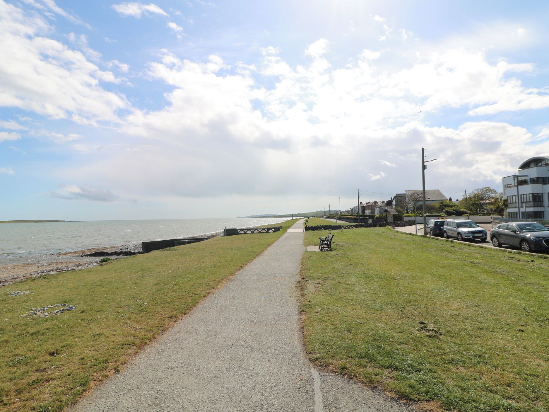 A coastal walkway with a bench, a path leading to the sea, and parked cars along the side.