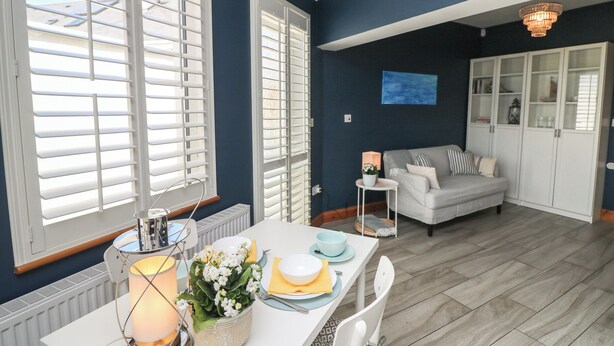 A dining area with a table set for two, a white sofa in the background, and a view of the outdoors through large windows with blinds. - To The Beach! (Skerries)