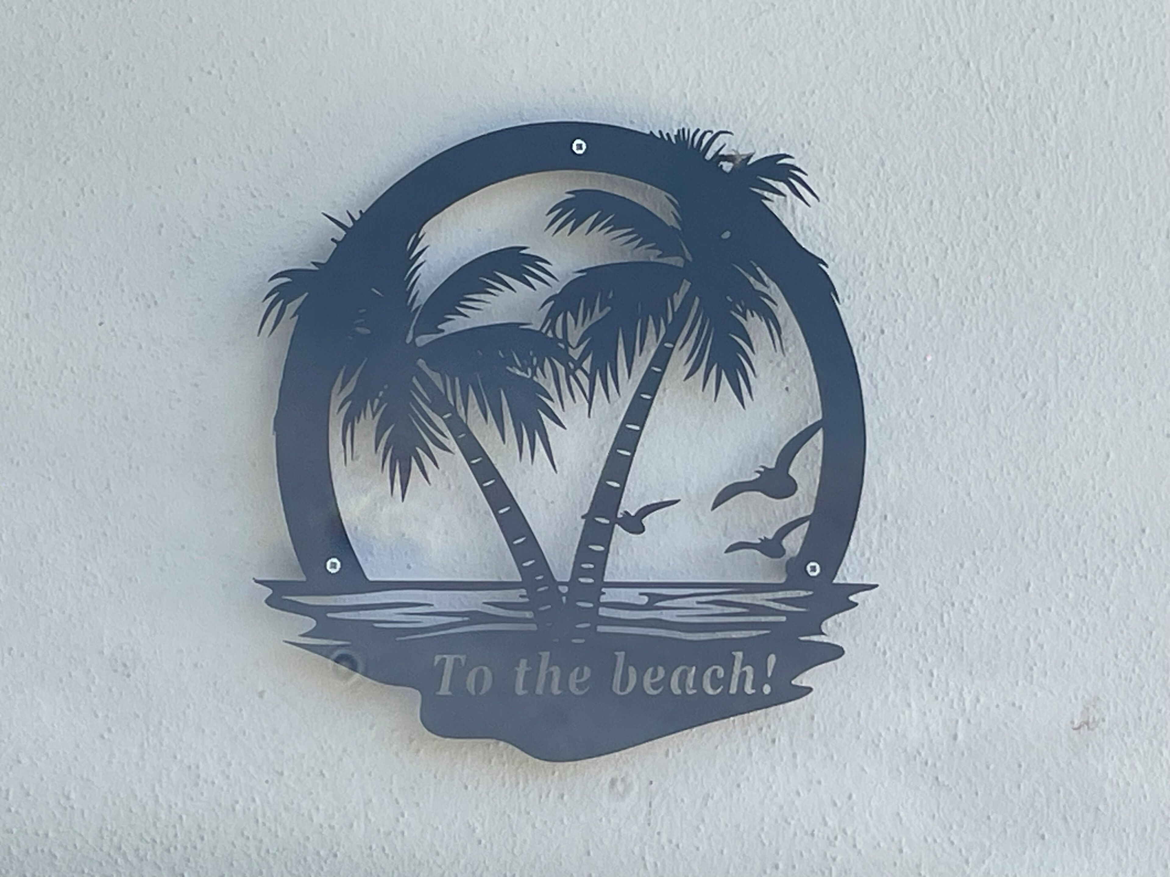 Une enseigne décorative en métal représentant des palmiers, des oiseaux et l’inscription : « À la plage ! »