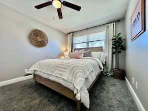 Een slaapkamer met een bed, een plafondventilator, een raam met gordijnen, een decoratief object aan de muur, en een potplant.