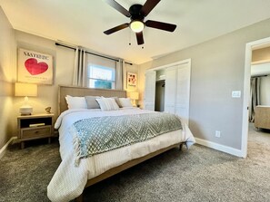 Ein Schlafzimmer mit Bett, Nachttisch, Deckenventilator, Fenster mit Vorhängen, einer Tür und einer Wand mit gerahmten Bildern.
