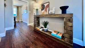 A hallway with a wooden floor, a built-in shelf with books and decorative items, a lamp, and a wall-mounted artwork. - All the Vibes Poolside in Trenton! (Trenton)