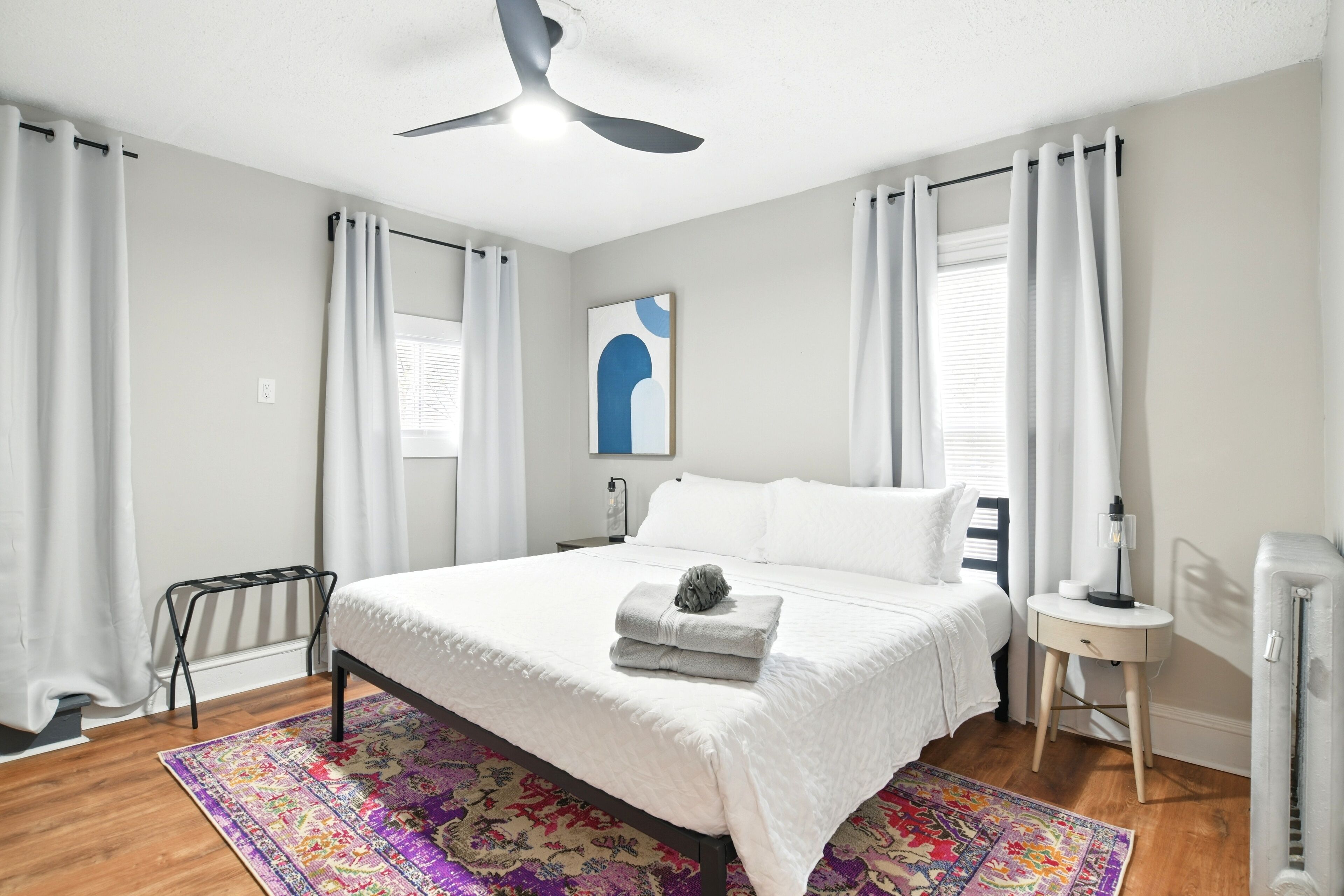 Un dormitorio ordenado con cama, un calefactor portátil, una mesita de noche pequeña y un ventilador de techo.